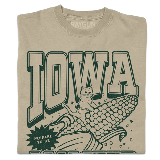 Tan garment-dyed t-shirt featuring a dark green graphic of a cat riding a giant ear of Iowa corn with the text IOWA and PREPARE TO BE.