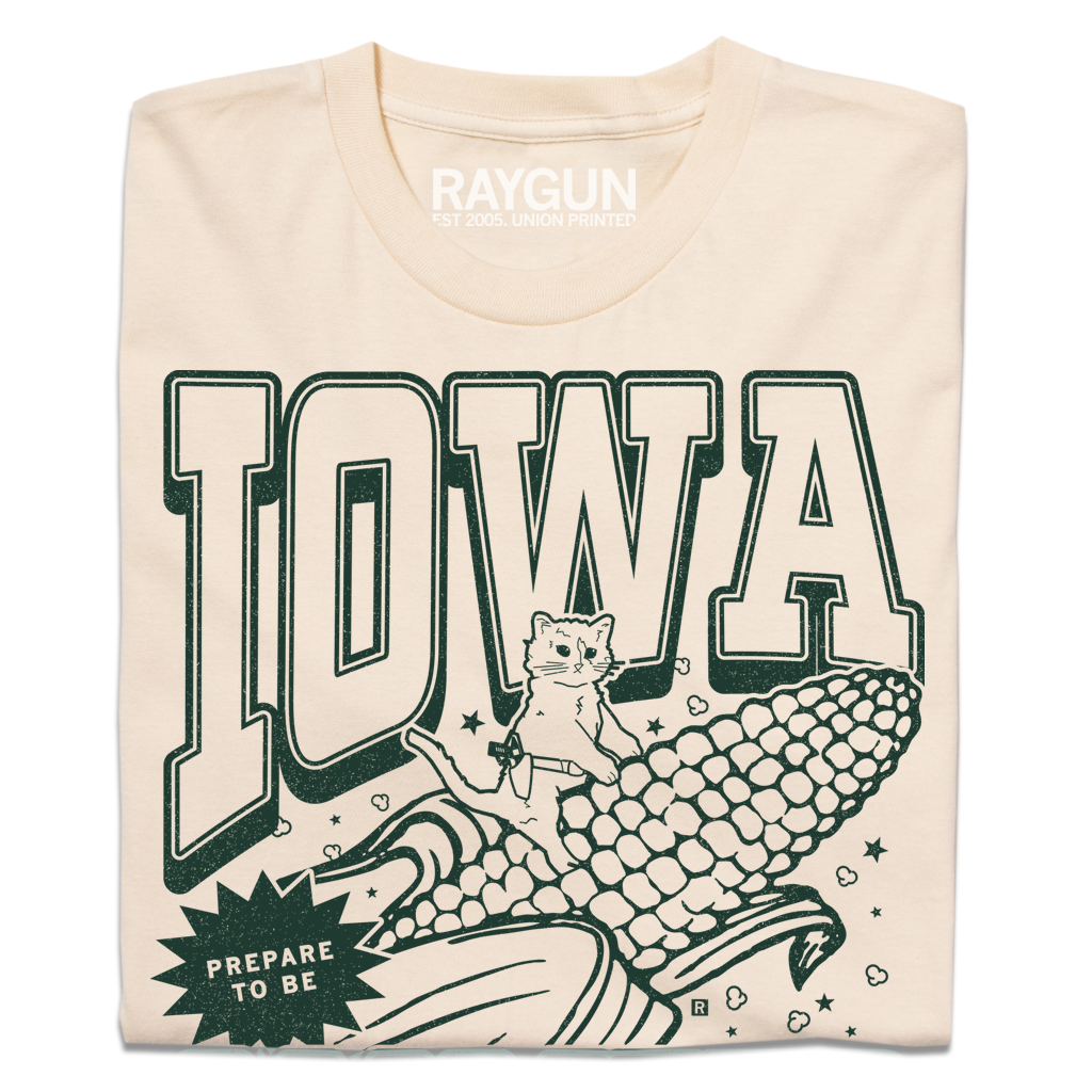 A folded cream t-shirt with dark green ink that says, "Iowa: Prepare to be aMAIZEd by all this f*cking corn" along with an illustration of a cat riding a giant ear of corn.