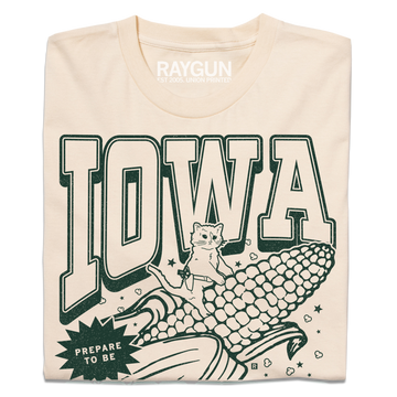 A folded cream t-shirt with dark green ink that says, "Iowa: Prepare to be aMAIZEd by all this f*cking corn" along with an illustration of a cat riding a giant ear of corn.