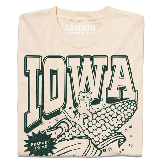A folded cream t-shirt with dark green ink that says, "Iowa: Prepare to be aMAIZEd by all this f*cking corn" along with an illustration of a cat riding a giant ear of corn.