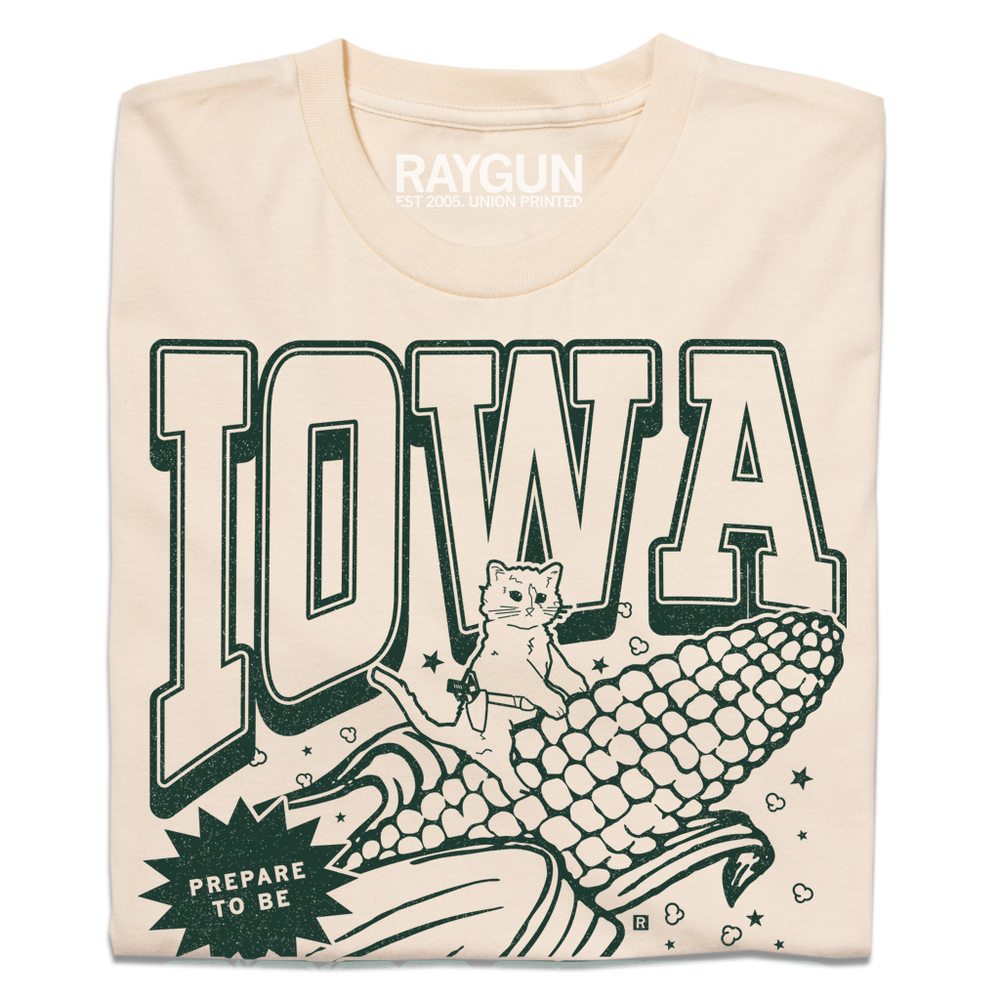 A folded cream t-shirt with dark green ink that says, "Iowa: Prepare to be aMAIZEd by all this f*cking corn" along with an illustration of a cat riding a giant ear of corn.