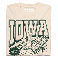 A folded cream t-shirt with dark green ink that says, "Iowa: Prepare to be aMAIZEd by all this f*cking corn" along with an illustration of a cat riding a giant ear of corn.