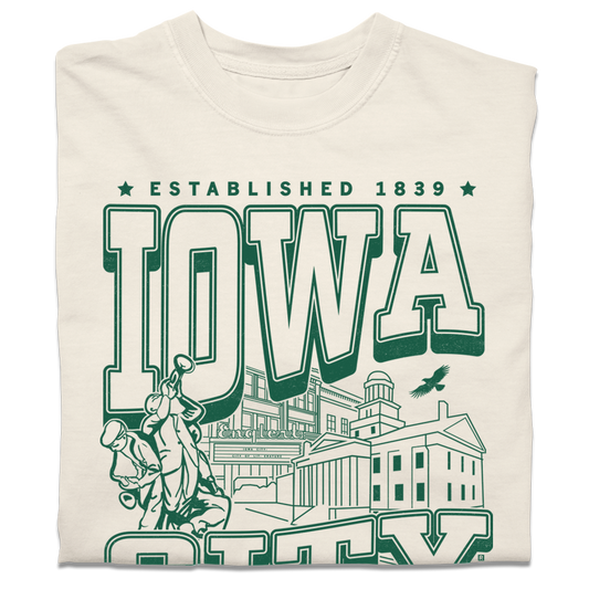 A folded heavyweight cream t-shirt with kelly green ink that says, "Iowa City: Established 1839. Iowa's first state Capital" with and illustrated Iowa City staples in the center, including the Englert Theater and the old Capitol Building.