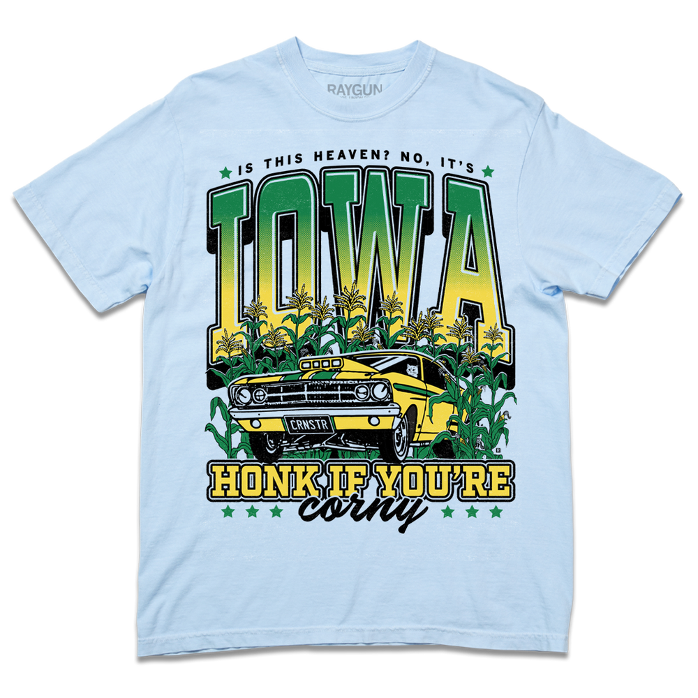 A Comfort Colors hydrangea t-shirt with green & gold ink that says, "Is this heaven? No, It's Iowa. Honk if you're corny" with a graphic of an old muscle car driving out of corn fields with a cat behind the steering wheel.