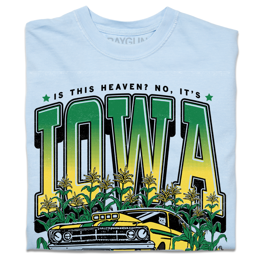 A folded heavyweight light blue t-shirt with green & gold ink that says, "Is this heaven? No, It's Iowa. Honk if you're corny" with a graphic of an old muscle car driving out of corn fields with a cat behind the steering wheel.