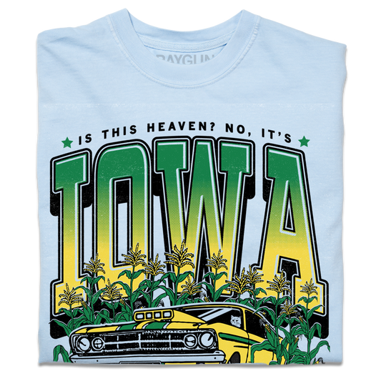 A folded heavyweight light blue t-shirt with green & gold ink that says, "Is this heaven? No, It's Iowa. Honk if you're corny" with a graphic of an old muscle car driving out of corn fields with a cat behind the steering wheel.
