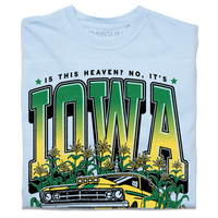 A folded heavyweight light blue t-shirt with green & gold ink that says, "Is this heaven? No, It's Iowa. Honk if you're corny" with a graphic of an old muscle car driving out of corn fields with a cat behind the steering wheel.
