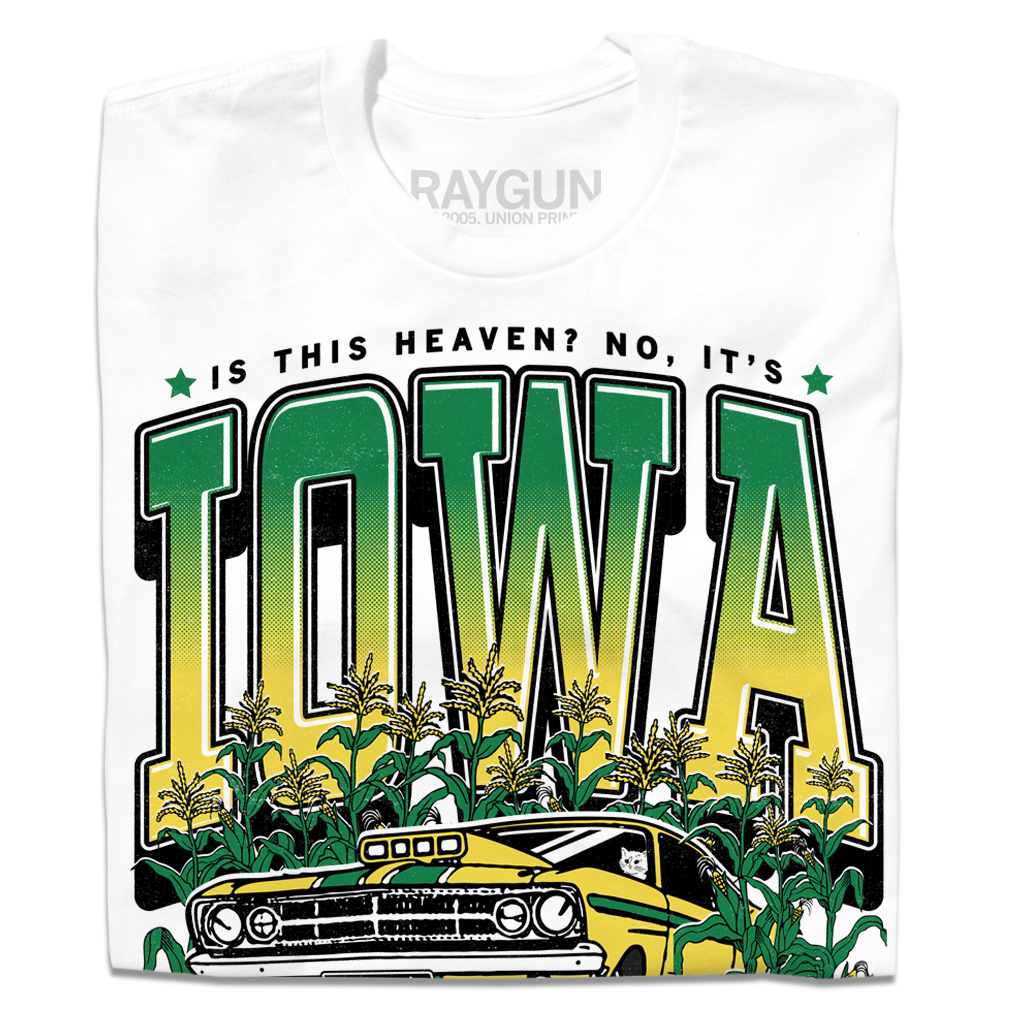 A folded white t-shirt with green & gold ink that says, "Is this heaven? No, It's Iowa. Honk if you're corny" with a graphic of an old muscle car driving out of corn fields with a cat behind the steering wheel.