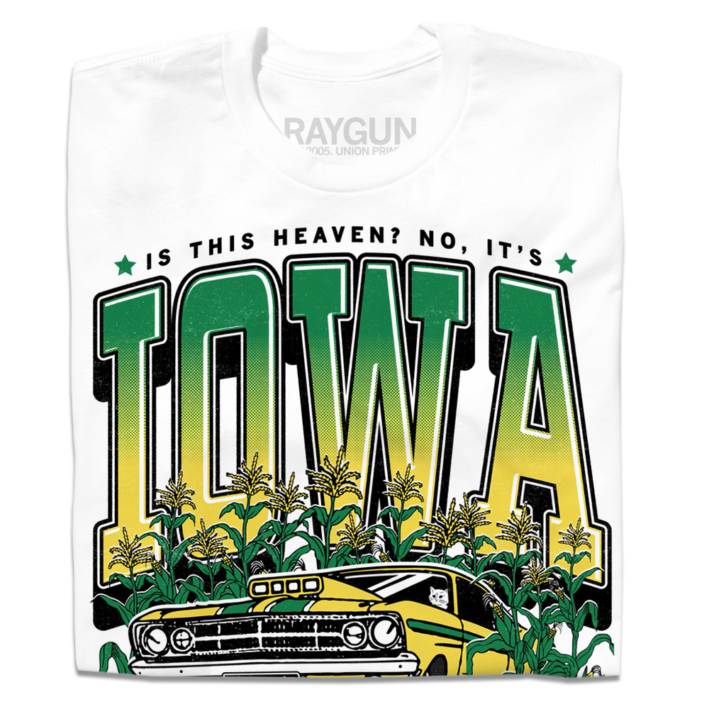 A folded white t-shirt with green & gold ink that says, "Is this heaven? No, It's Iowa. Honk if you're corny" with a graphic of an old muscle car driving out of corn fields with a cat behind the steering wheel.