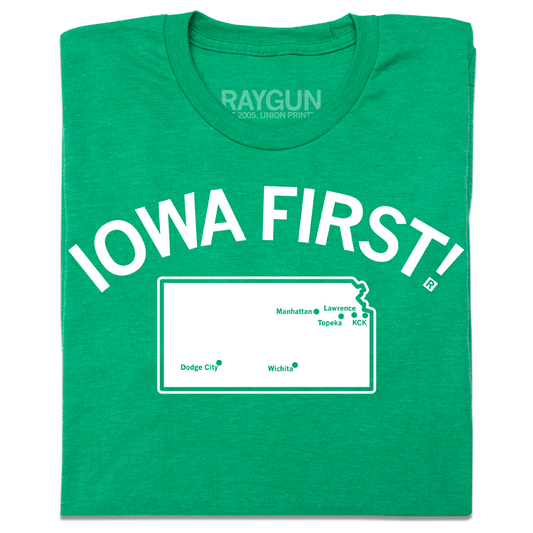 A folded heather kelly green t-shirt with white ink print. Curved text near the top says, "IOWA FIRST!" above a map of Kansas with cities in KS marked on it like a map.