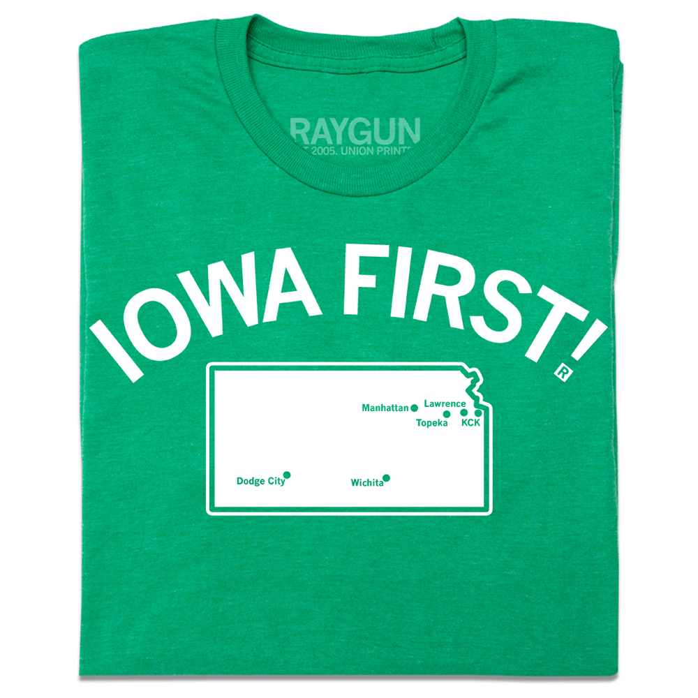 A folded heather kelly green t-shirt with white ink print. Curved text near the top says, "IOWA FIRST!" above a map of Kansas with cities in KS marked on it like a map.
