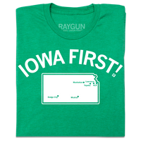 A folded heather kelly green t-shirt with white ink print. Curved text near the top says, "IOWA FIRST!" above a map of Kansas with cities in KS marked on it like a map.