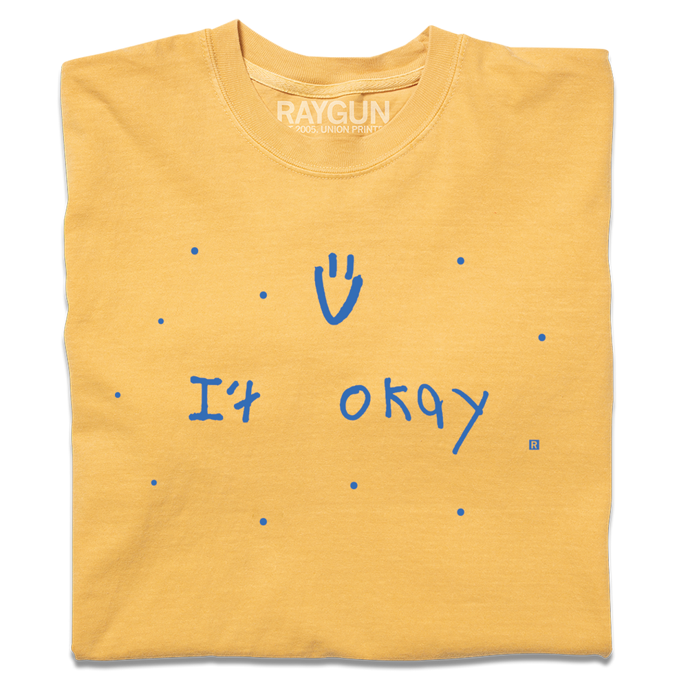 A folded heavyweight yellow t-shirt with blue print that says, "It Okay" with a smiley face above and dots scattered around. This shirt was designed by a kid taking part in a contest at our Cedar Rapids store.