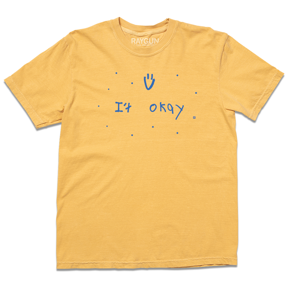 Comfort Colors heavyweight mustard t-shirt with blue print that says, "It Okay" with a smiley face above and dots scattered around. This shirt was designed by a kid taking part in a contest at our Cedar Rapids store.