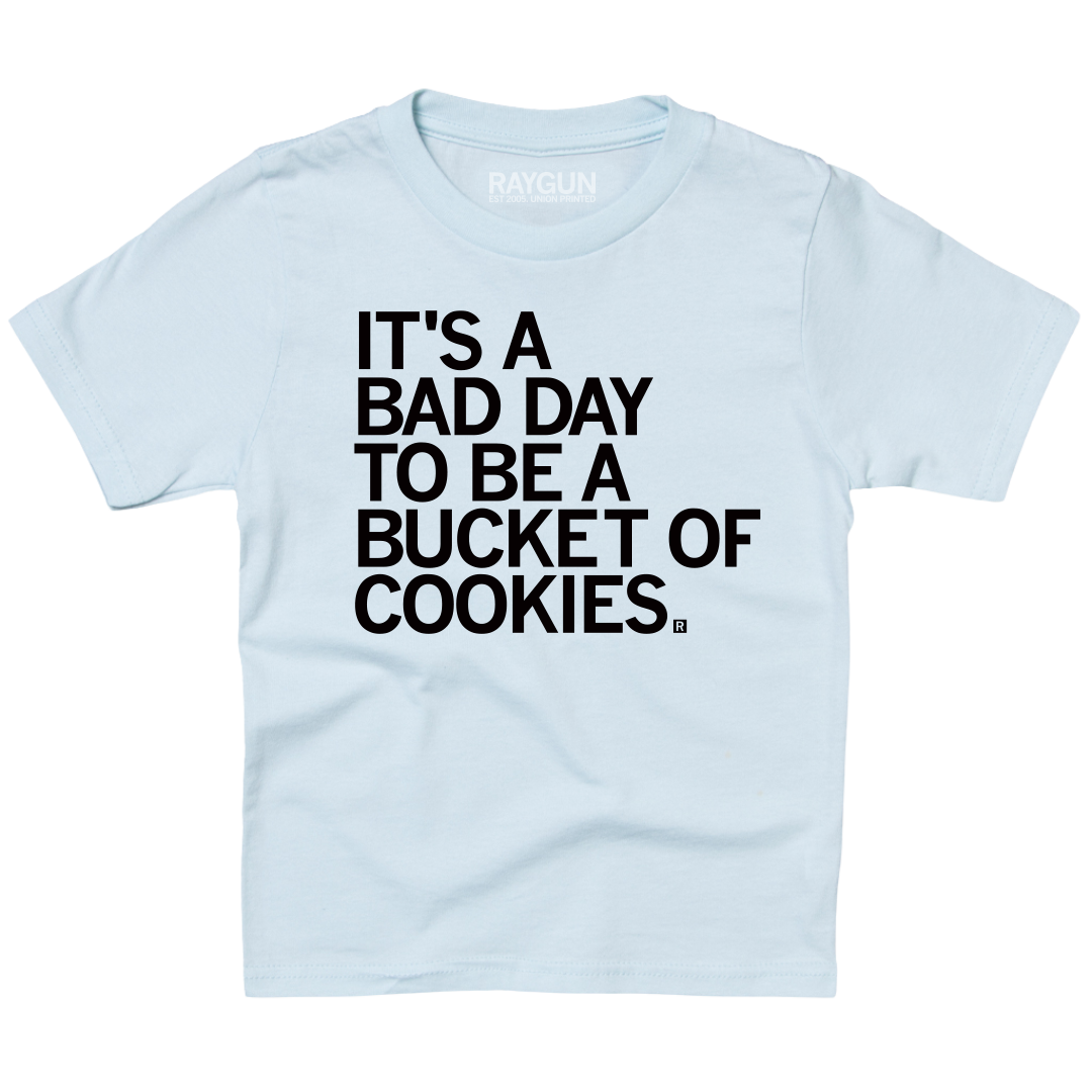 A light blue youth sized t-shirt with left aligned black text in all caps that says, "It's a bad day to be a bucket of cookies."