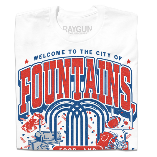 White shirt with red & blue ink that says, "Welcome to the city of fountains, food, and football. Kansas City, Missouri USA" with a fountain, football, helmet, goalpost, smoker, and steak in the background.