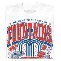 White shirt with red & blue ink that says, "Welcome to the city of fountains, food, and football. Kansas City, Missouri USA" with a fountain, football, helmet, goalpost, smoker, and steak in the background.