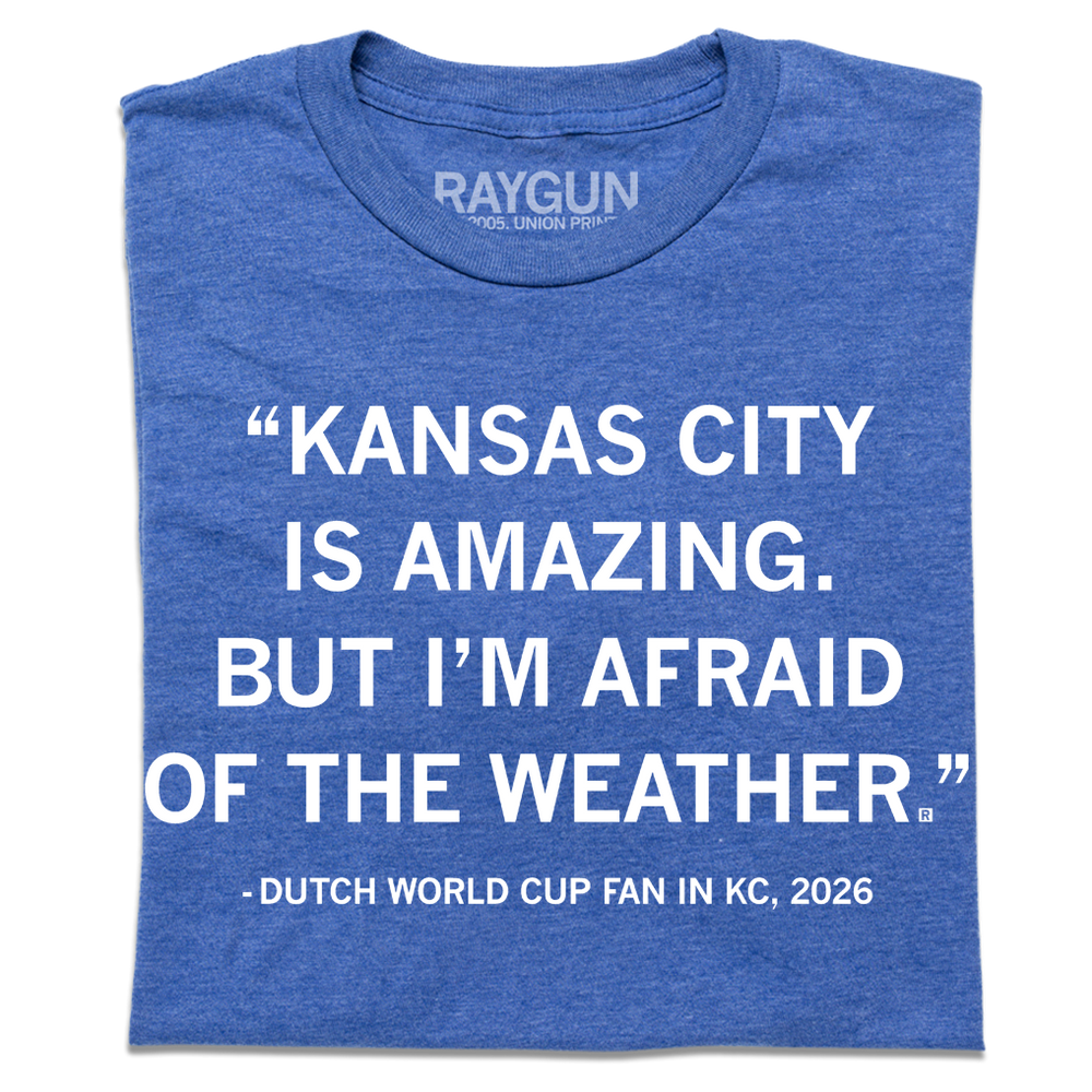 A folded heather blue t-shirt with white ink text that says, "Kansas City is amazing, but I'm afraid of the weather." - Dutch World Cup Fan in KC, 2026."