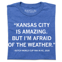 A folded heather blue t-shirt with white ink text that says, "Kansas City is amazing, but I'm afraid of the weather." - Dutch World Cup Fan in KC, 2026."
