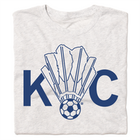 A folded heather white shirt with royal blue ink depicting a shuttlecock with a soccer ball at the end of it between the letters KC.