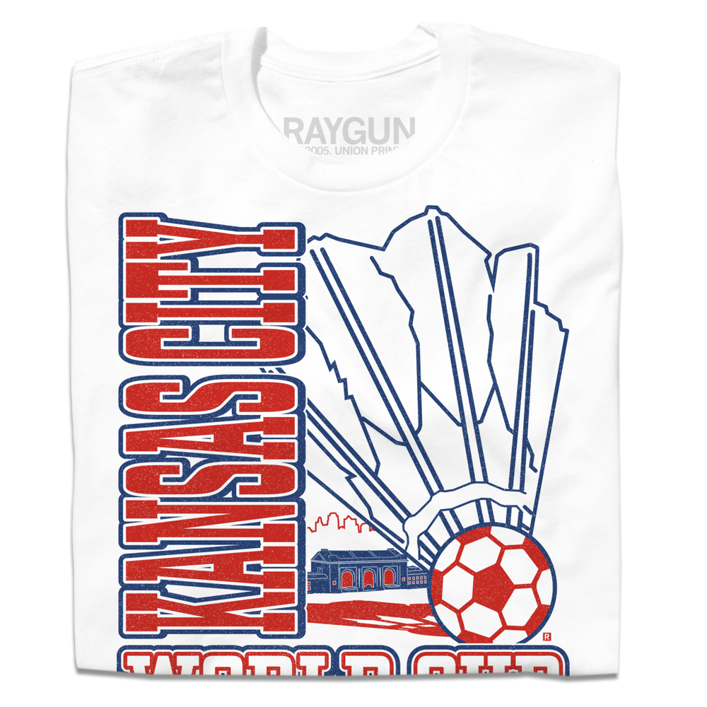 A folded white t-shirt with red & royal blue ink depicting a giant shuttlecock with a soccer ball at the end in front of an outline of the KC skyline. Text reads "World Cup Kansas City, Missouri. USA 2026."