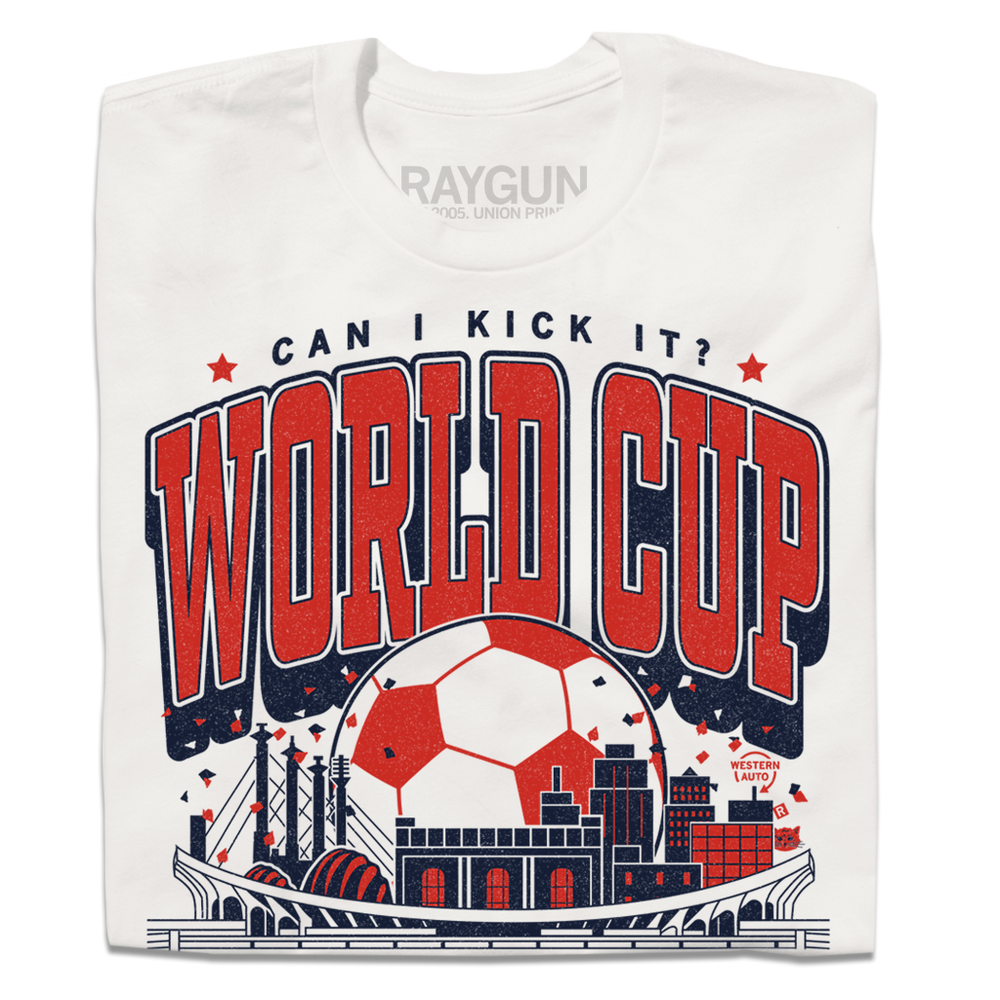 A folded white t-shirt with red & navy ink depicting notable buildings around Kansas City behind a soccer stadium and in front of a giant soccer ball. Text reads "Can I kick it? World Cup Kansas City, Missouri. USA 2026."