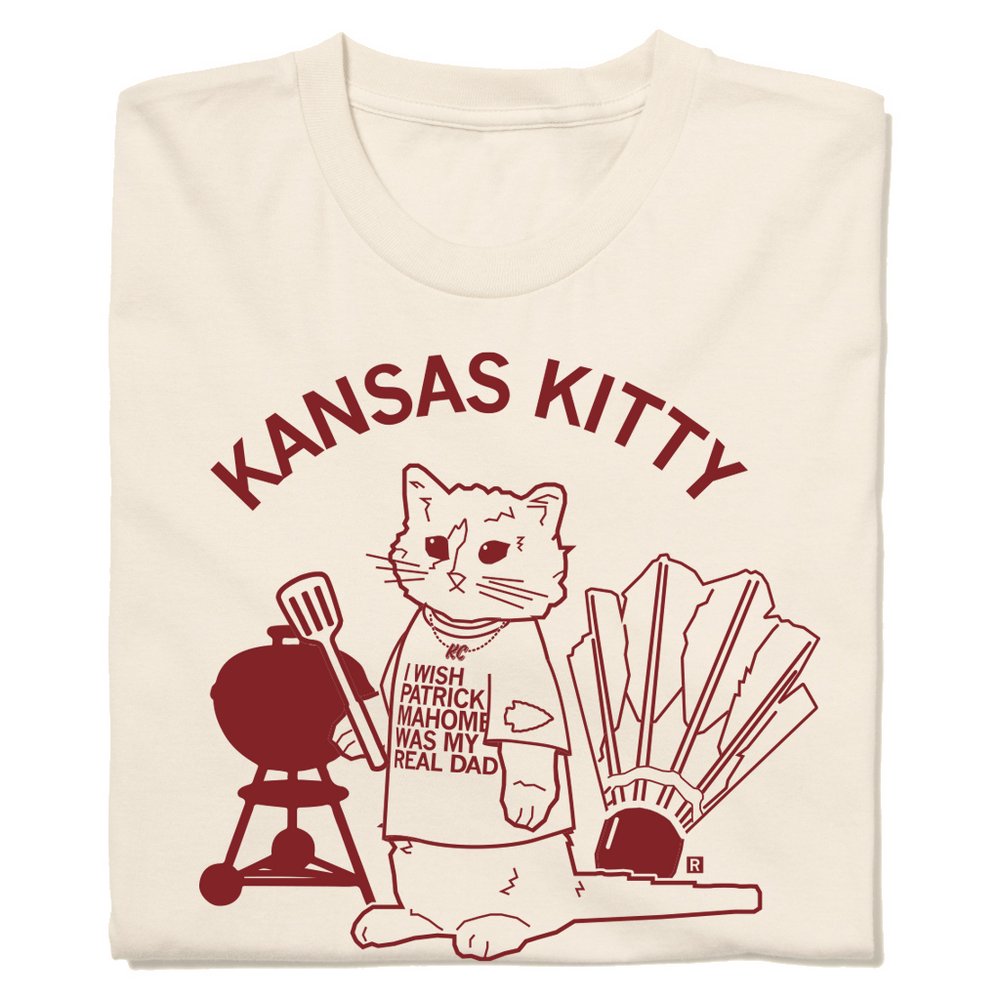 A folded off white t-shirt with an image of RAYGUN's cat mascot Garry wearing a shirt that says, "I wish Patrick Mahomes Was My Real Dad" while standing between a grill and a shuttlecock. In cardinal text above this reads "Kansas Kitty"