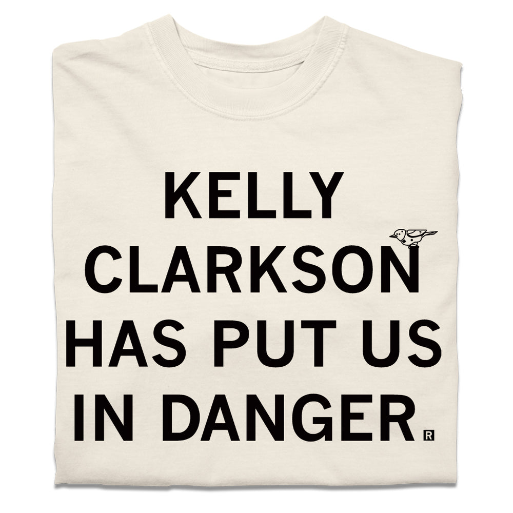 A folded cream heavyweight t-shirt with blank ink text that says, "Kelly Clarkson has put us in danger." with a little bird sitting on the N in "Clarkson."