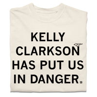 A folded cream heavyweight t-shirt with blank ink text that says, "Kelly Clarkson has put us in danger." with a little bird sitting on the N in "Clarkson."