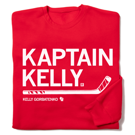 A folded red crewneck sweatshirt with white ink print that says "Kaptain Kelly" in all caps above a horizontal hockey stick. Smaller text below adds "Kelly Gorbatenko" beside a tiny outline of Wisconsin with the number 7 in it.