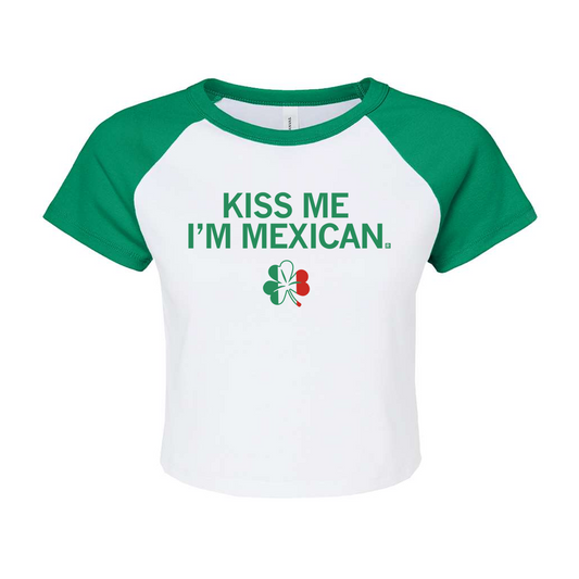 White cropped baby tee with green raglan sleeves, featuring the text KISS ME IM MEXICAN above a clover icon colored with the Mexican flag.