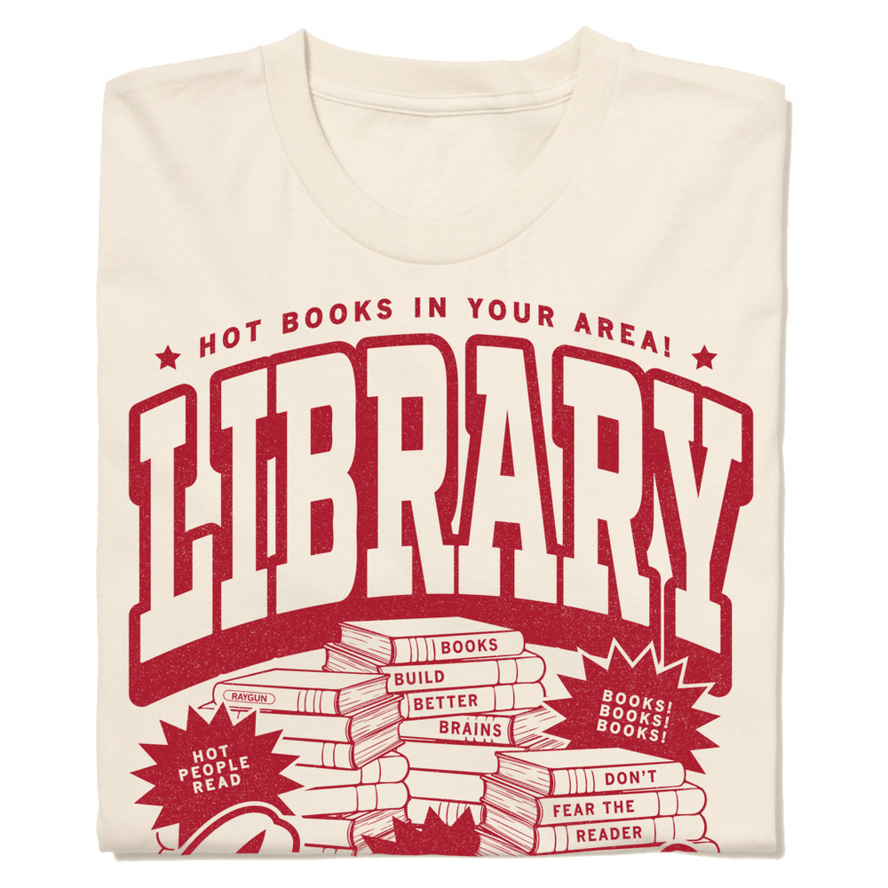 Off white t-shirt with cardinal ink. Small font across the top reads "Hot Books in your area!" then large, outlined text says, "Library Lover" with an illustration of piles of books in the center.