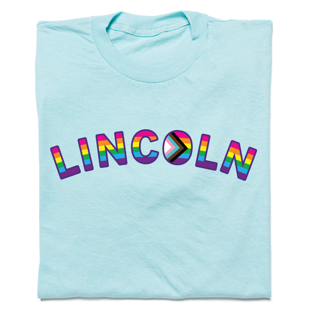 Light blue RAYGUN t-shirt featuring the word LINCOLN in rainbow-colored letters, with the letter O designed as a Progress Pride flag for Lincoln NE Pride.
