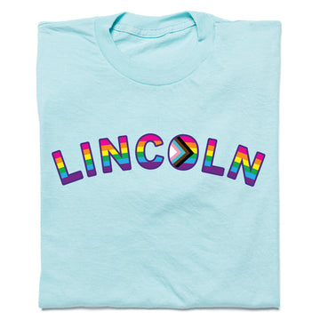 Light blue RAYGUN t-shirt featuring the word LINCOLN in rainbow-colored letters, with the letter O designed as a Progress Pride flag for Lincoln NE Pride.