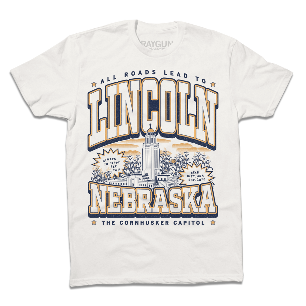 A Next Level Bone t-shirt with vintage gold and navy ink depicting the Nebraska Capitol in the center. Small text at the top reads "All roads lead to" then large curved text says, "Lincoln" above the capitol and "Nebraska" below it. Small text at the very bottoms reads "the cornhusker capitol."