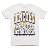 A Next Level Bone t-shirt with vintage gold and navy ink depicting the Nebraska Capitol in the center. Small text at the top reads "All roads lead to" then large curved text says, "Lincoln" above the capitol and "Nebraska" below it. Small text at the very bottoms reads "the cornhusker capitol."