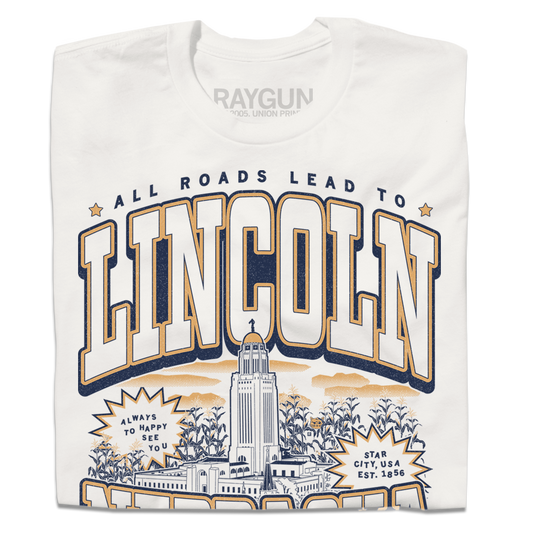 A folded dark white t-shirt with vintage gold and navy ink depicting the Nebraska Capitol in the center. Small text at the top reads "All roads lead to" then large curved text says, "Lincoln" above the capitol and "Nebraska" below it. Small text at the very bottoms reads "the cornhusker capitol."