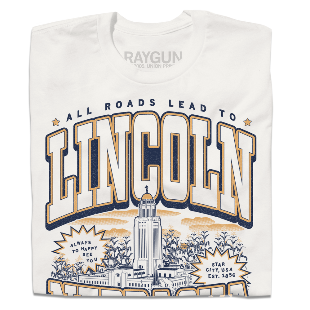 A folded dark white t-shirt with vintage gold and navy ink depicting the Nebraska Capitol in the center. Small text at the top reads "All roads lead to" then large curved text says, "Lincoln" above the capitol and "Nebraska" below it. Small text at the very bottoms reads "the cornhusker capitol."