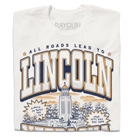 A folded dark white t-shirt with vintage gold and navy ink depicting the Nebraska Capitol in the center. Small text at the top reads "All roads lead to" then large curved text says, "Lincoln" above the capitol and "Nebraska" below it. Small text at the very bottoms reads "the cornhusker capitol."