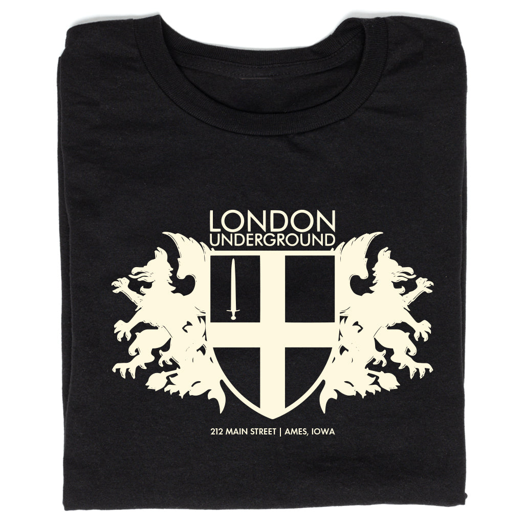 Black RAYGUN t-shirt featuring a white London Underground crest logo with two griffins flanking a sword-bearing shield, with text below noting 212 MAIN STREET | AMES, IOWA.