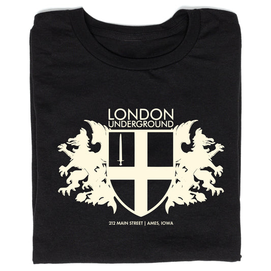 Black RAYGUN t-shirt featuring a white London Underground crest logo with two griffins flanking a sword-bearing shield, with text below noting 212 MAIN STREET | AMES, IOWA.