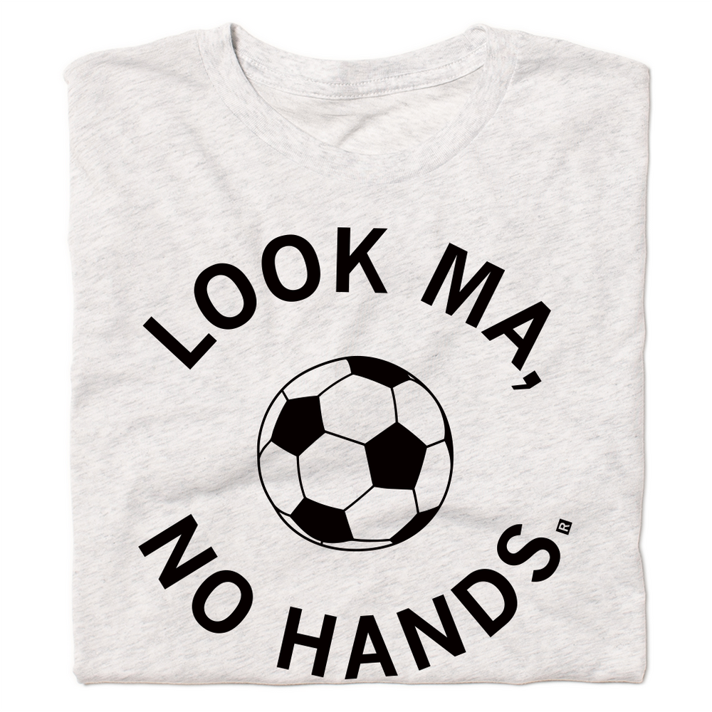 A folded heather white t-shirt with black ink. An illustrated soccer ball in the center is surrounded by curved text that reads "Look ma, no hands."