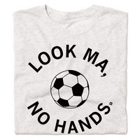 A folded heather white t-shirt with black ink. An illustrated soccer ball in the center is surrounded by curved text that reads "Look ma, no hands."