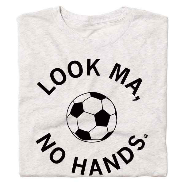 A folded heather white t-shirt with black ink. An illustrated soccer ball in the center is surrounded by curved text that reads 