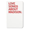 A white RAYGUN notebook cover featuring the red text LOVE SONGS ABOUT MADISON.