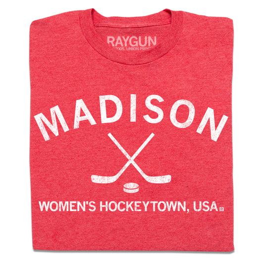 A folded heather red t-shirt with white ink that reads "Madison. Women's Hockeytown, USA." with crossing hockey sticks and a puck in the center.