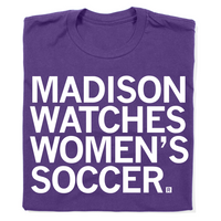 A folded purple t-shirt with left aligned text in white ink that reads "Madison watches women's soccer"