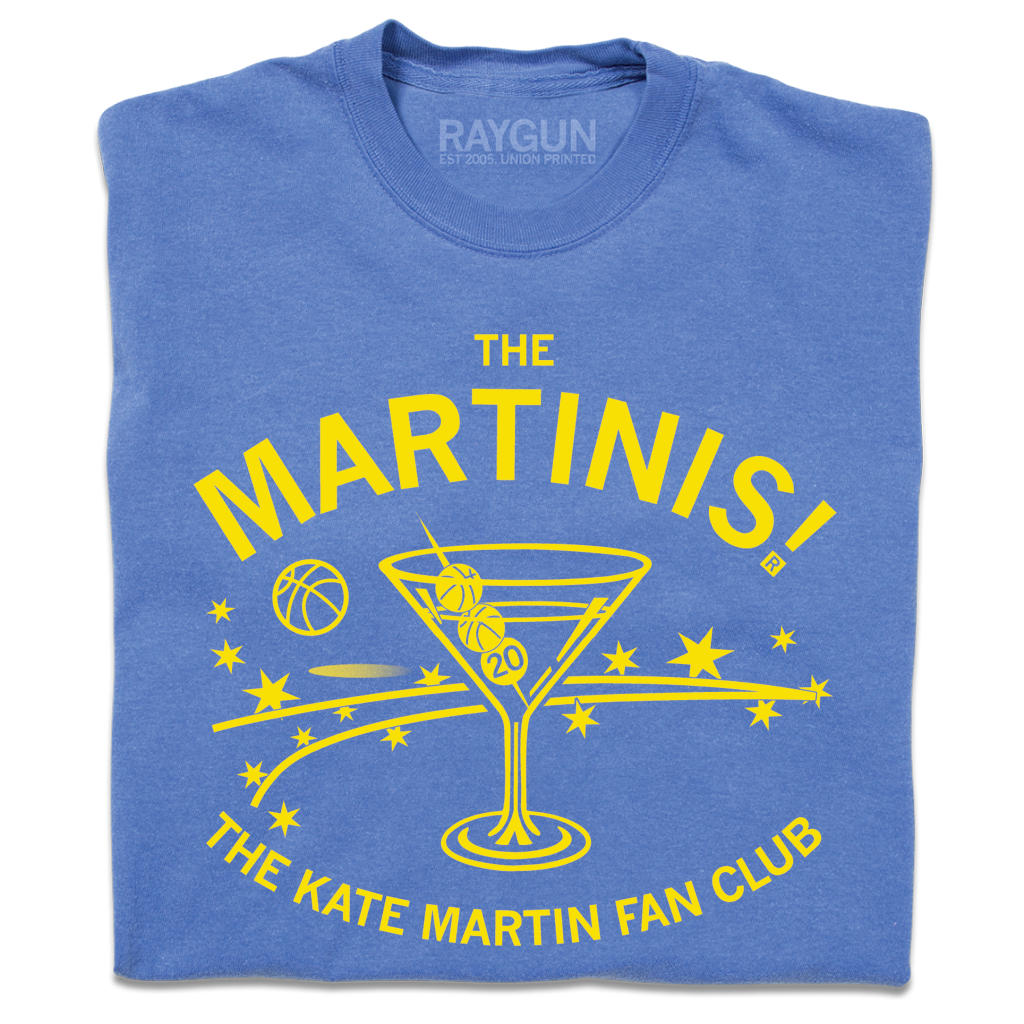 A folded blue heavyweight t-shirt with bright yellow ink. The center is an illustration of a martini glass with tiny basketballs on a toothpick in place of olives. Curved text above says "The MARTINIS!" and smaller fond below the image adds "THE KATE MARTIN FAN CLUB." There's also a bouncing basketballs, scatters stars, and other accents.