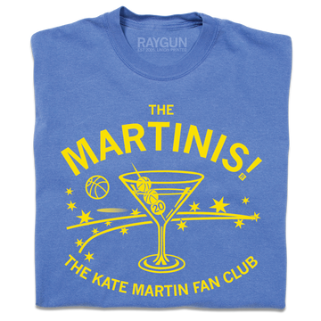 A folded blue heavyweight t-shirt with bright yellow ink. The center is an illustration of a martini glass with tiny basketballs on a toothpick in place of olives. Curved text above says "The MARTINIS!" and smaller fond below the image adds "THE KATE MARTIN FAN CLUB." There's also a bouncing basketballs, scatters stars, and other accents.