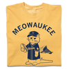 A folded mustard yellow t-shirt with navy ink that says, "MEOWAUKEE" in curved text over Gary The Cat wearing Brewers gear while holding a hotdog.